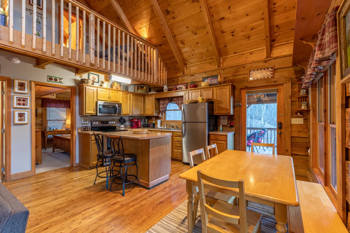 This image captures the inviting dining area and kitchen, with wooden beams above, a kitchen island, a rustic dining table, and large windows that offer scenic mountain views. The setup suggests a space for family meals, morning coffee, or casual gatherings, with open sightlines that foster connection between the kitchen and dining areas.