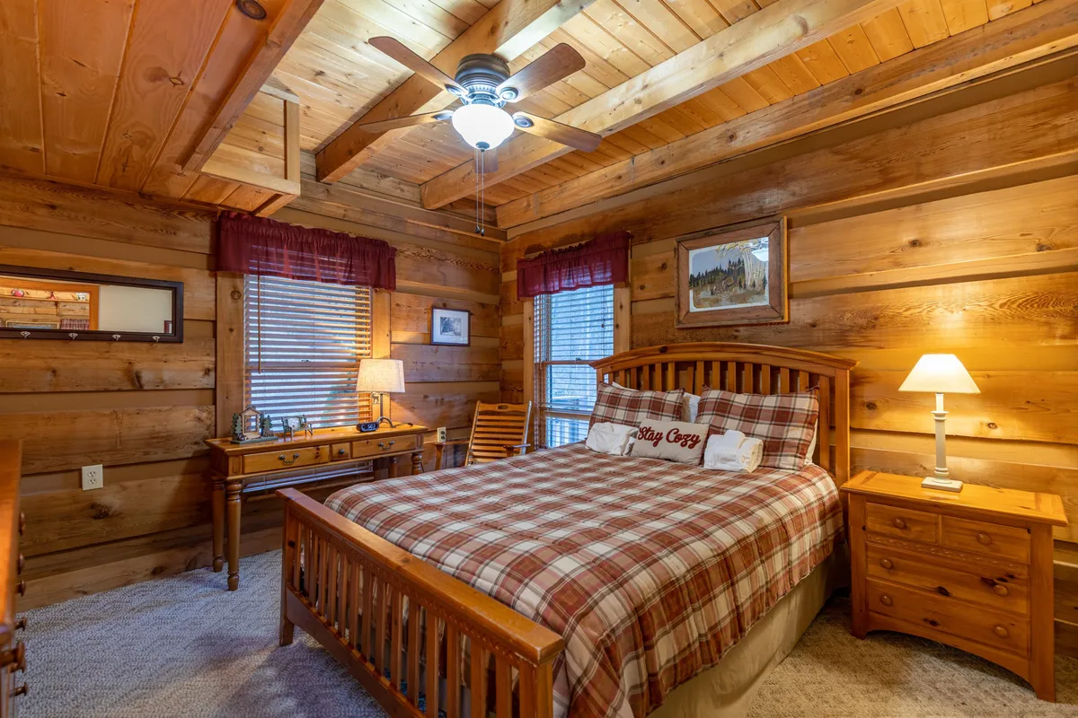 A cozy bedroom featuring a wooden bed frame topped with plaid bedding, a ceiling fan overhead, and a small writing desk with a lamp by the window. The room feels warm and inviting, ideal for unwinding after a day outdoors.