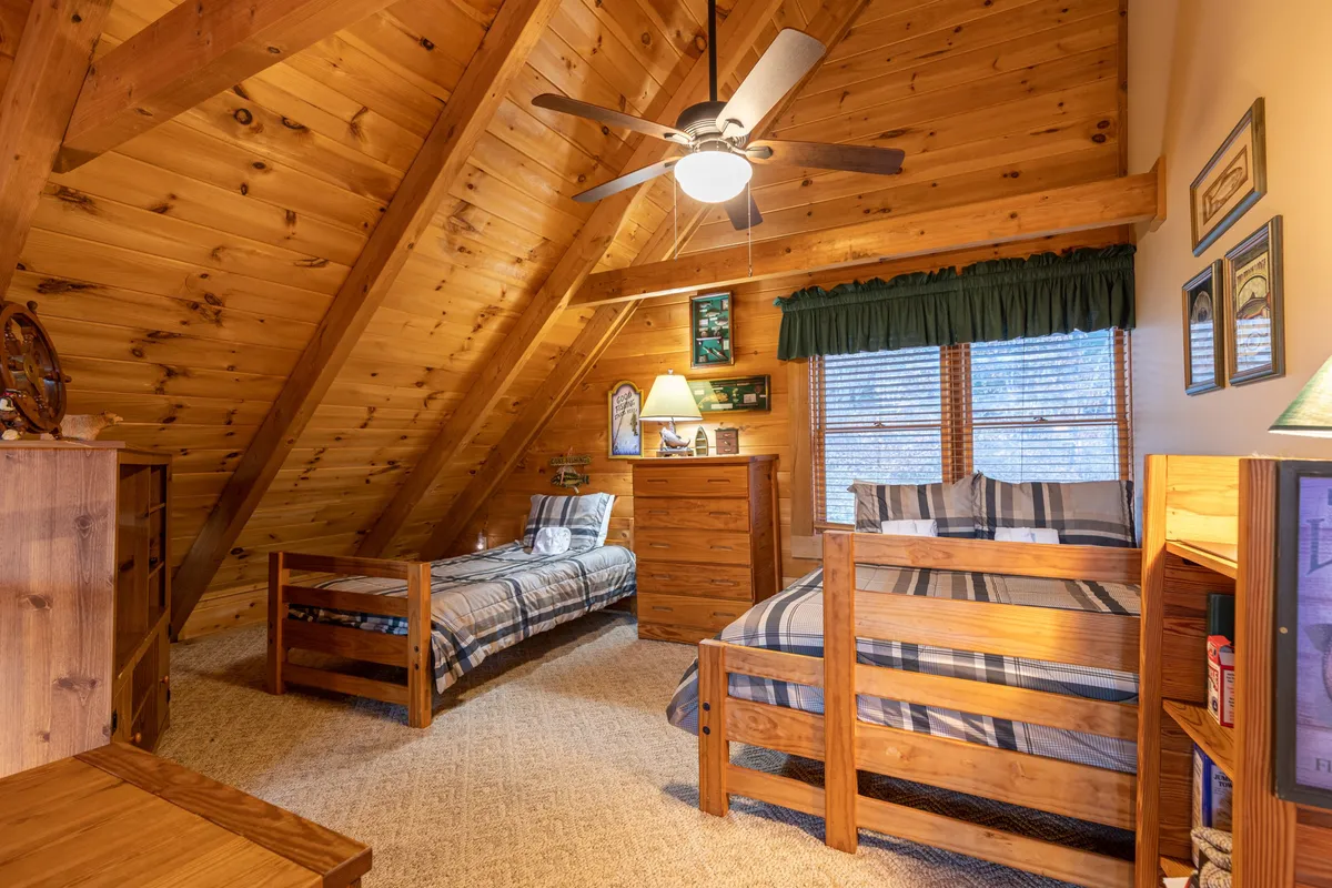 A charming loft area with wooden beams, cozy bunk beds dressed in soft blankets, a chest of drawers, a lamp, and a window with blinds letting in natural light. The space is both functional and inviting.