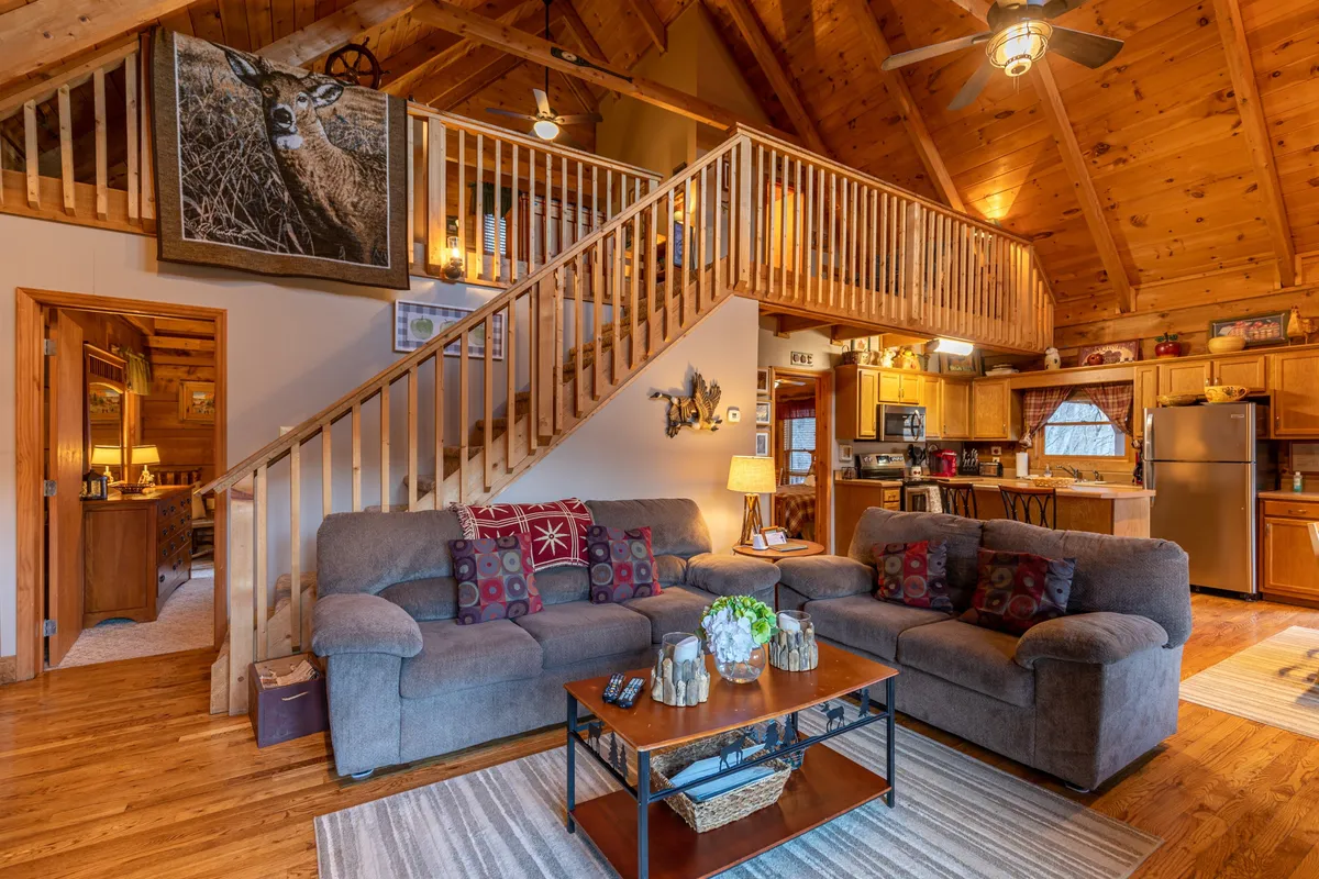 A cozy, open-plan living area featuring a plush sofa, coffee table, and a view of the adjacent kitchen area. The staircase and wall art add a homey touch, while the space is arranged for socializing and relaxation. The inviting ambiance is perfect for family gatherings or group activities like playing games or sharing stories.