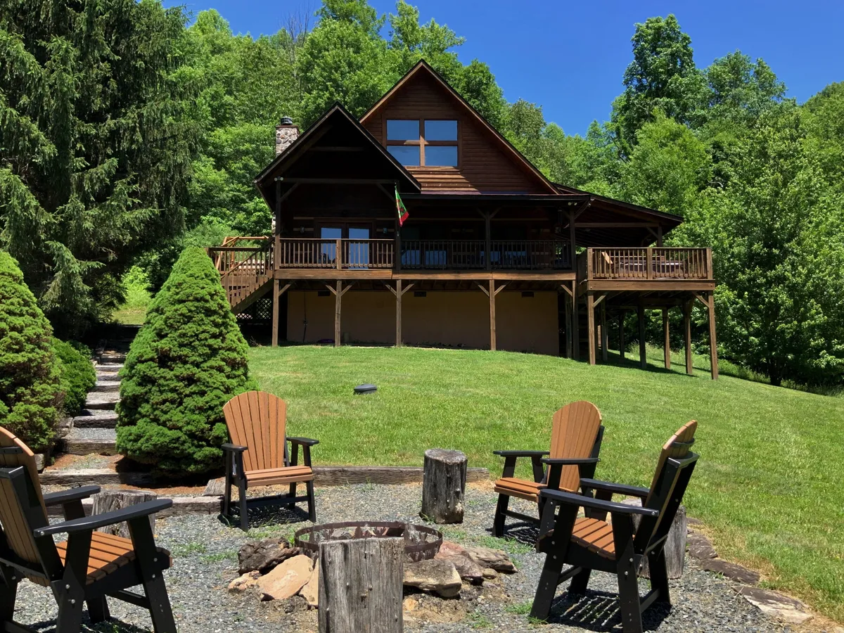 The hero image shows a charming log cabin surrounded by trees, with a spacious deck, inviting fire pit, and a cozy outdoor seating area overlooking a peaceful pasture and pond. Adirondack chairs circle the fire pit, perfect for gatherings under the stars, and a side deck offers a spot for grilling and sunset views.