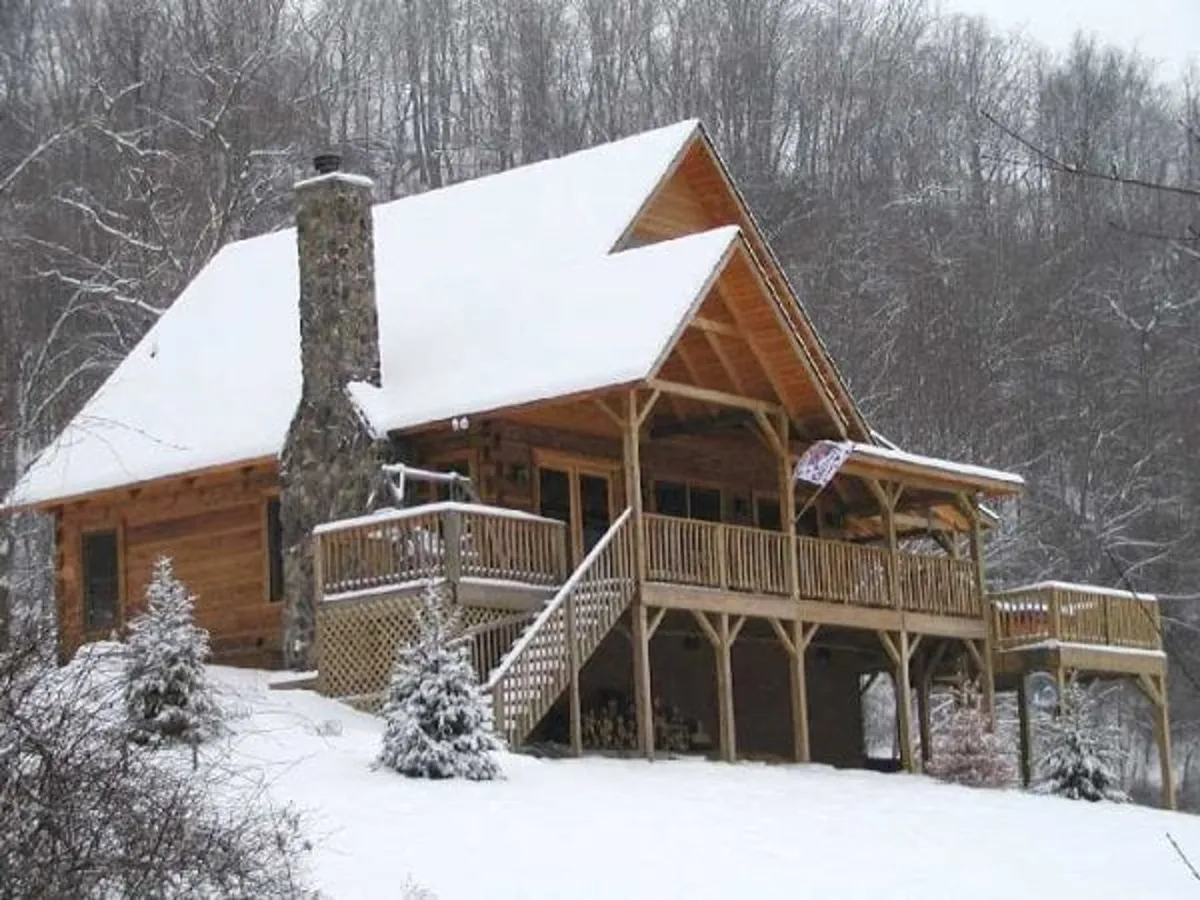 This image captures the exterior of the wooden cabin nestled in a snow-covered mountain landscape. The stone chimney and decorative railing add rustic charm, while the surrounding woods and thick blanket of snow evoke a sense of seclusion and adventure. The image hints at outdoor activities like snowball fights and evenings by the fire pit, as well as cozy moments sipping hot chocolate on the porch.