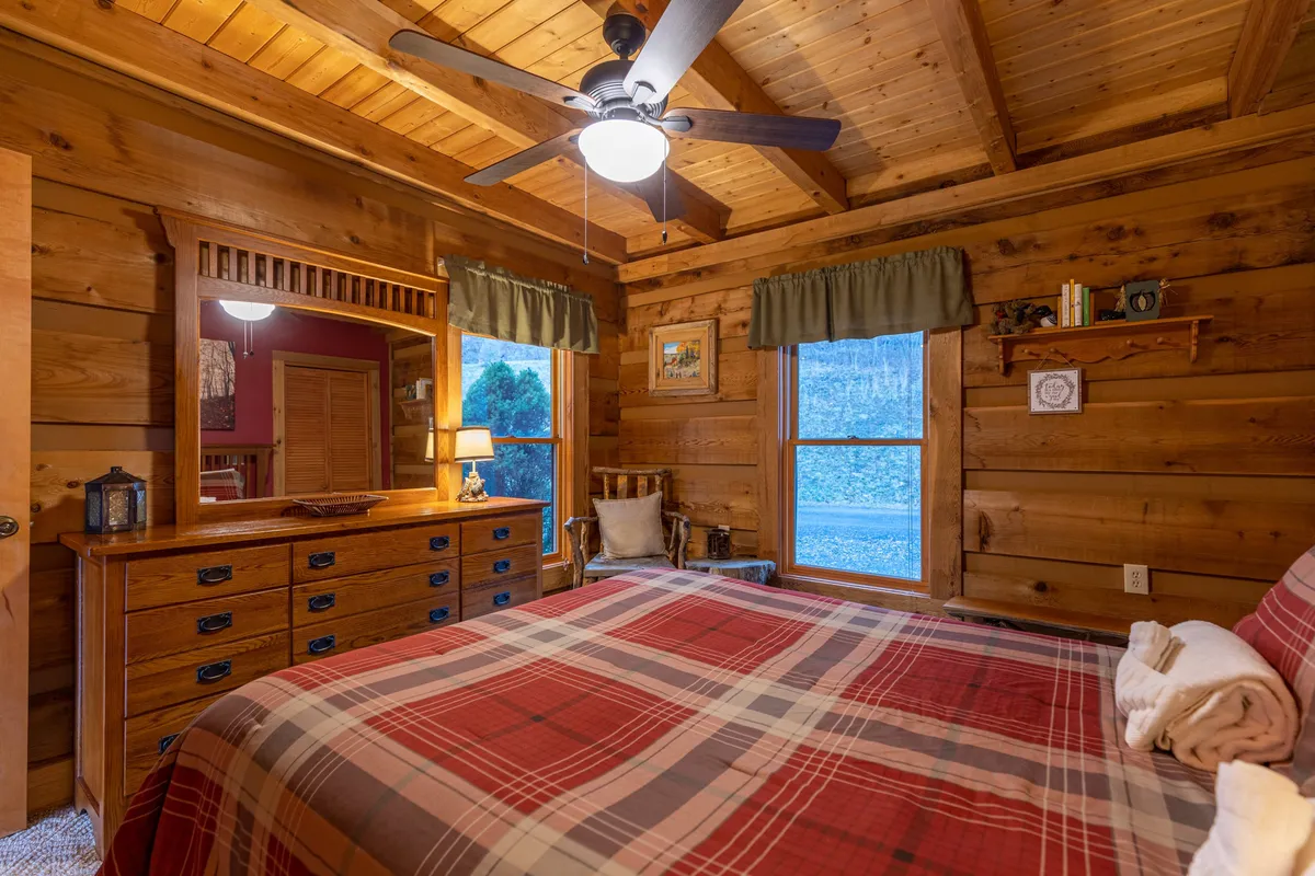 A cozy, warmly lit bedroom with a plaid bedspread, wooden dresser, and ceiling fan. Sunlight trickles through curtained windows, inviting a sense of comfort and relaxation. The inviting setting hints at restful evenings and intimate moments spent unwinding together after a day of exploration.