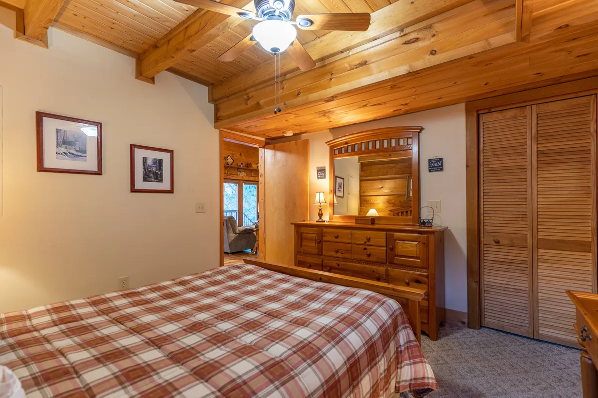 A second restful bedroom with a plaid bedspread, wooden furniture, a dresser with a mirror, decorative wall art, and a ceiling fan. The room captures soft natural light, creating a serene setting.