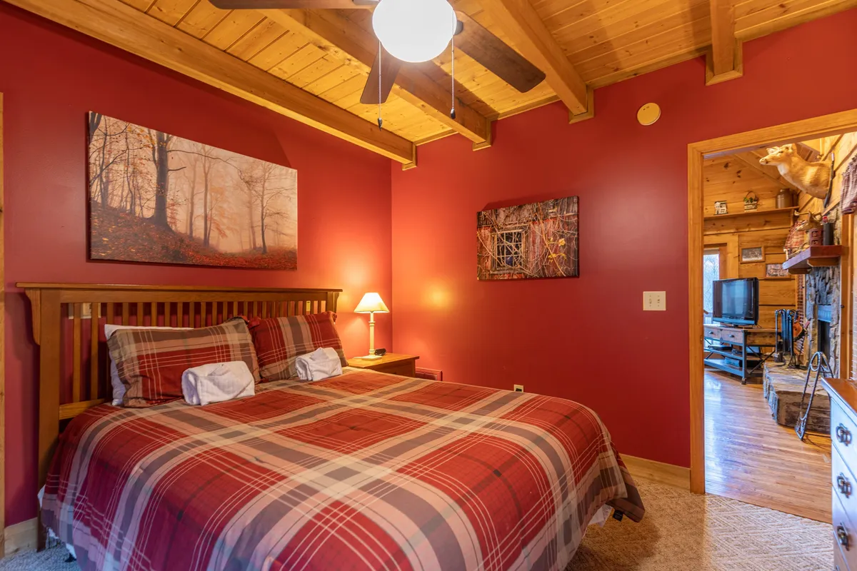 A tranquil bedroom scene featuring a bed with plaid bedding, rustic wooden ceiling beams, and soft lamp lighting. Artwork on the walls and a bedside table create a homey, peaceful ambiance, ideal for unwinding after adventuring outdoors.