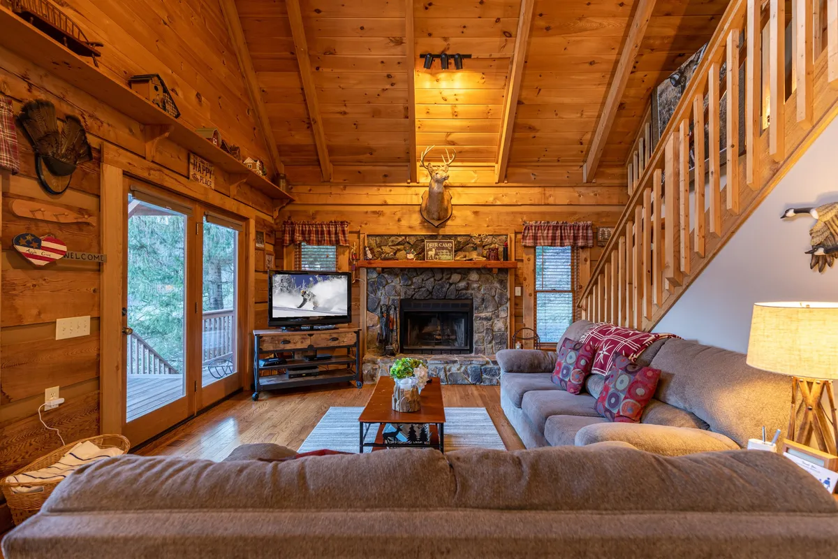 A welcoming living room featuring a stone fireplace, comfortable sofa, television, wooden beams, and decorative accents. Large windows bring in natural light and frame views of the outdoors, creating a cozy spot for family or friends to unwind during any season.
