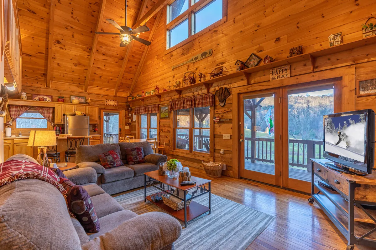This image showcases the main living area of the cabin, featuring a plush sofa, a television, wooden beams overhead, expansive windows that let in natural light, and a wood-burning fireplace as the central focal point. Decorative touches on the shelves and the cozy seating arrangement evoke a warm, communal atmosphere, perfect for family gatherings and movie nights.